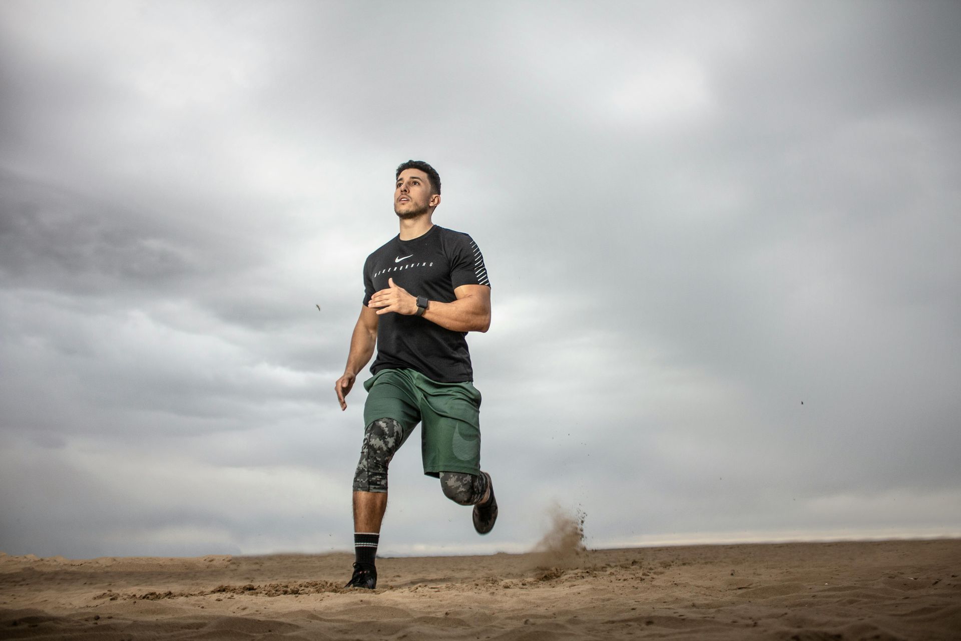 Man runs on sandy ground under a cloudy sky. He wears a black shirt, camo shorts, and black socks.