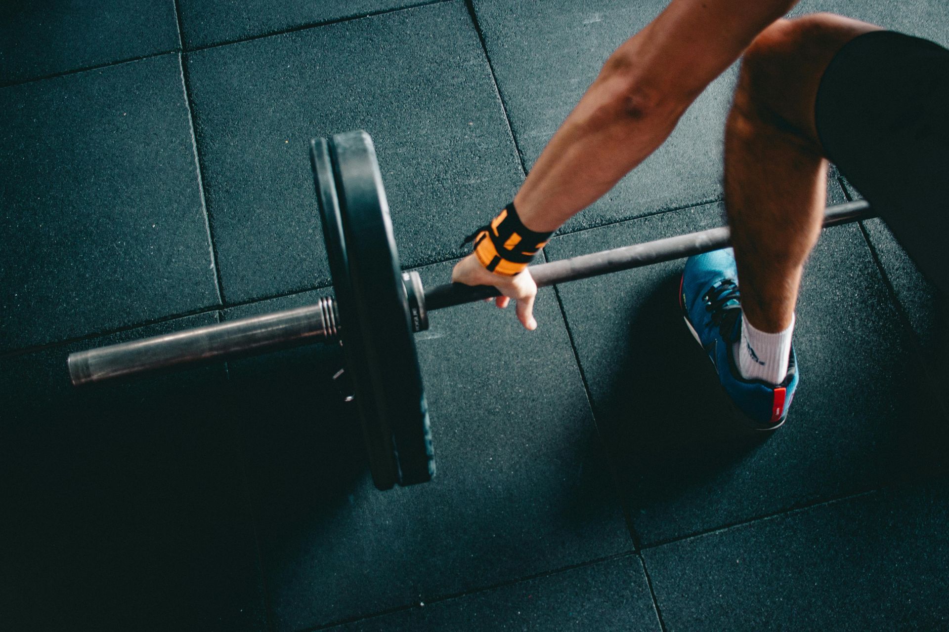 Person’s hand gripping a barbell in a gym, with a weight plate on the side. They're wearing wrist support and a blue shoe.