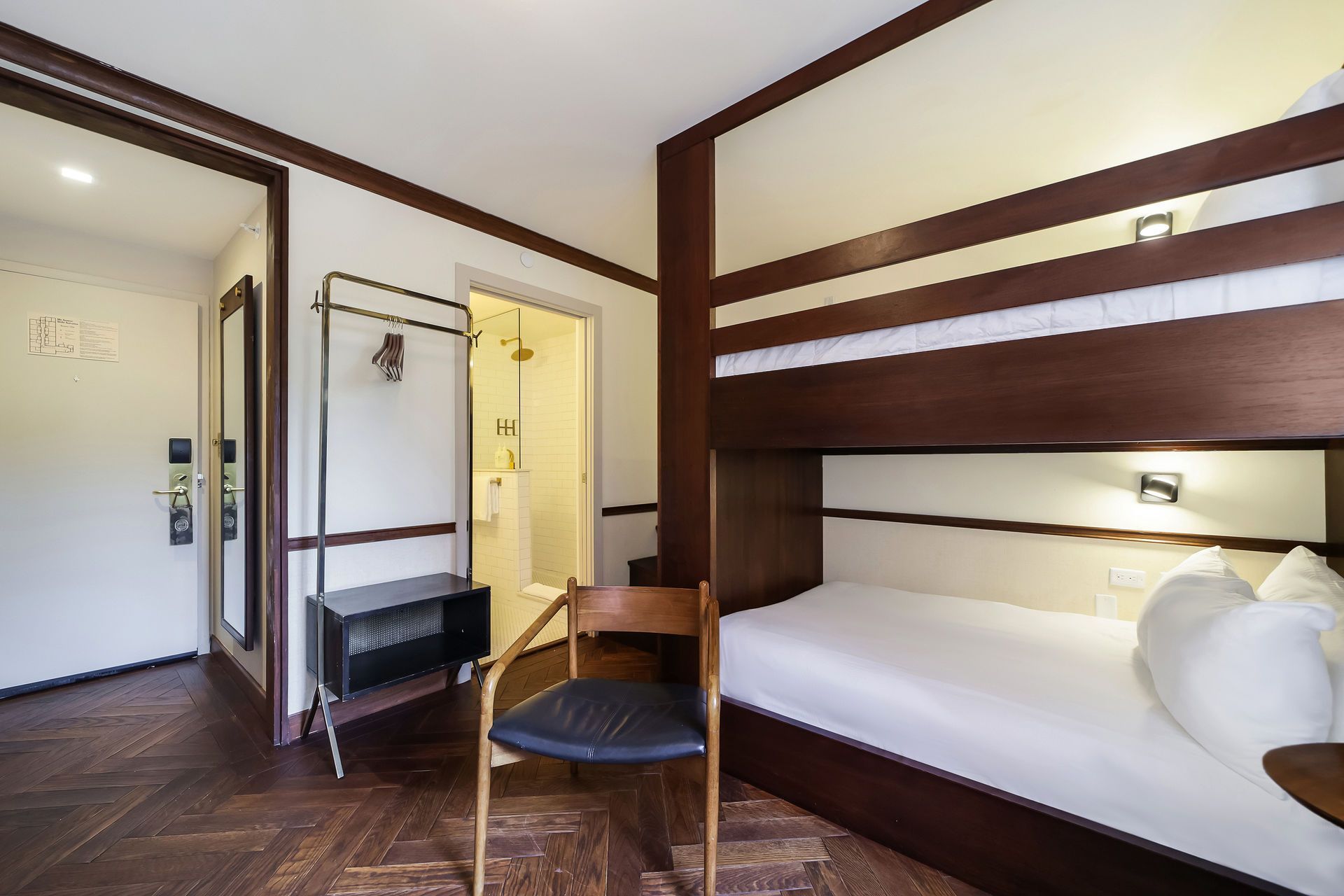 A modern hotel room with dark wood bunk beds, a wooden chair, a clothing rack, and a doorway leading to a bathroom.