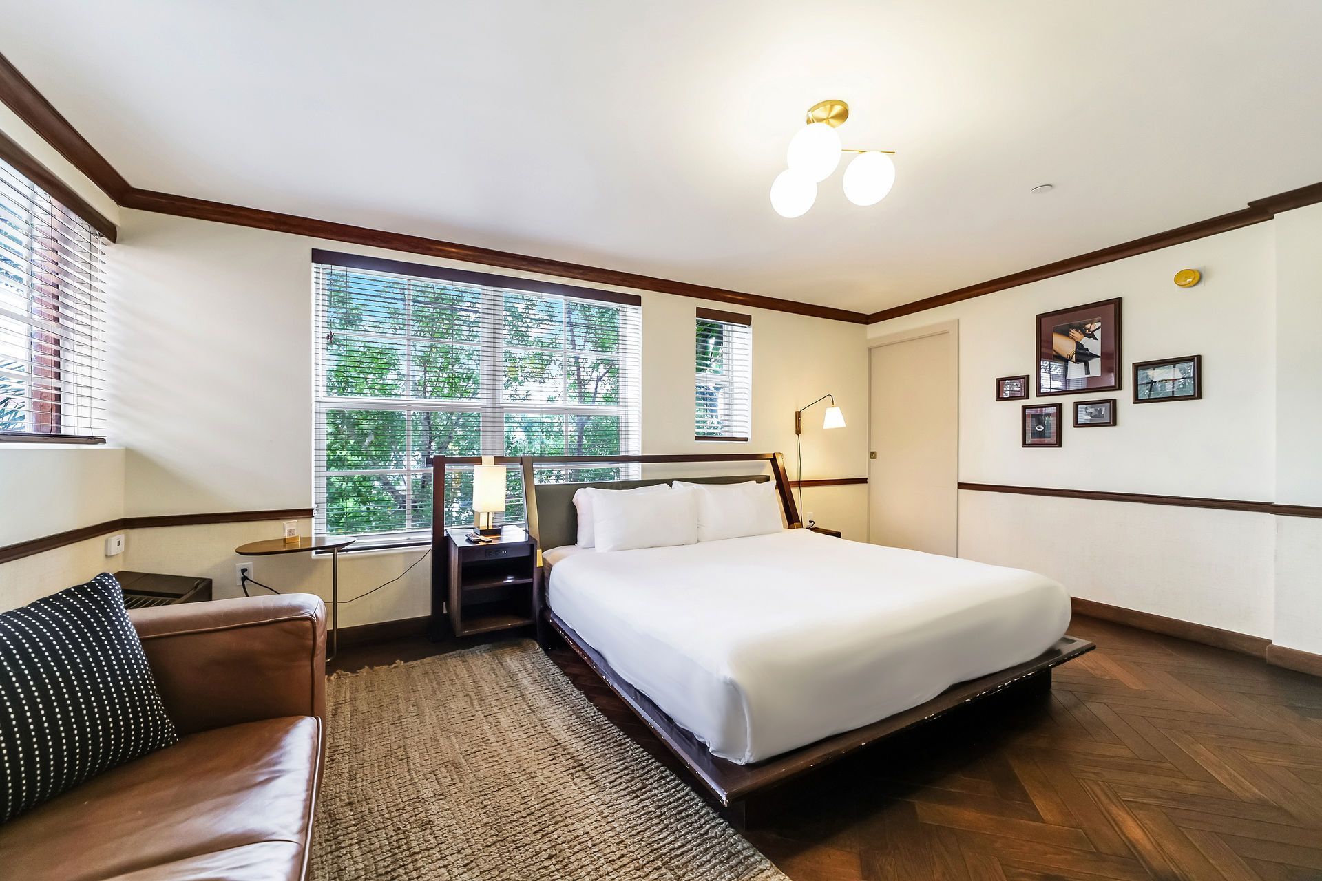 A modern bedroom with a large bed, brown leather sofa, wood floors, and photos on the wall under warm lighting.