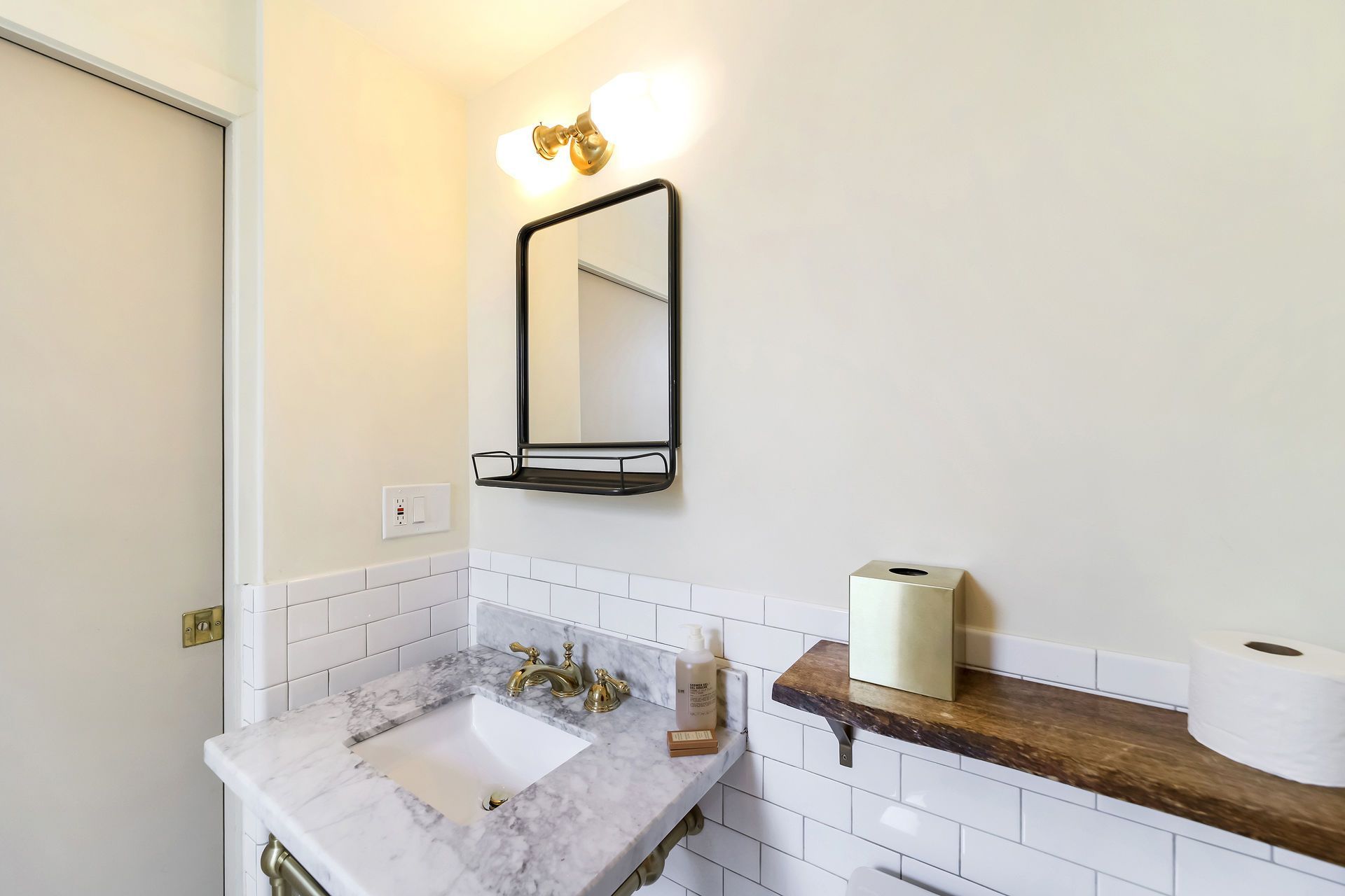 Bathroom with white tile, marble countertop, black mirror, and wood shelf holding tissue and a roll.
