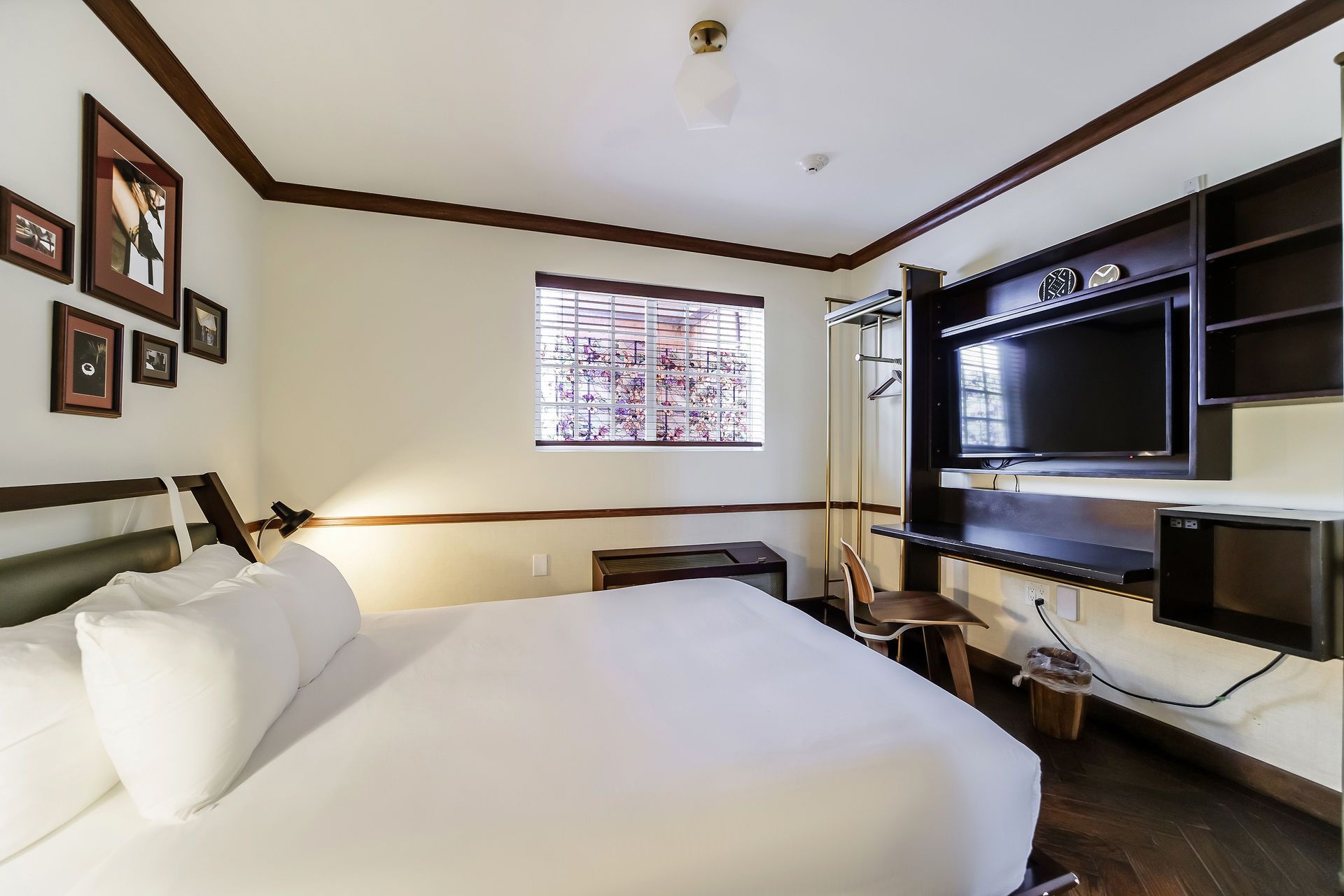 A bright hotel room featuring a white bed, dark wood furniture, a television, and a window with a view of greenery.