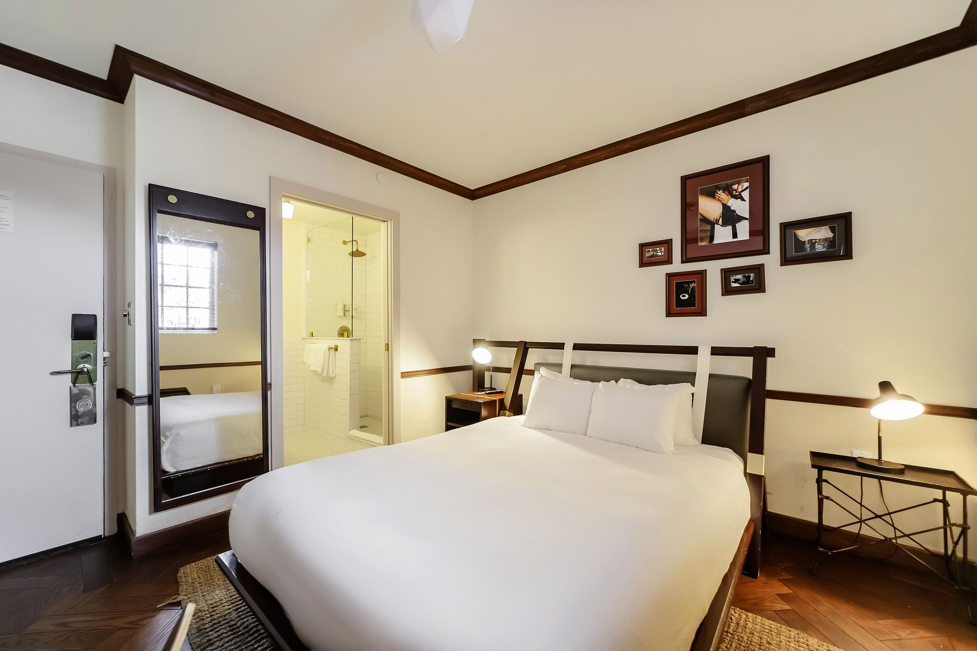 Hotel bedroom with a bed, small side tables, dark wood trim, and a shower visible through an open door.