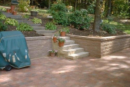 Brick patio with steps leading up a retaining wall to a landscaped yard. A grill is covered to the left.