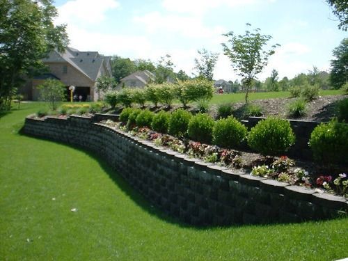 A curved retaining wall with green lawn, landscaping with shrubs and flowers, and a house in the background.
