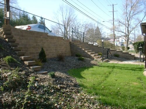 Stone retaining walls with steps leading up, a chain-link fence, and a grassy area.