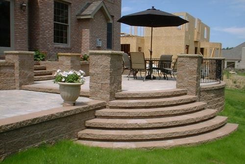 Stone patio with curved steps, table, chairs, and umbrella. Brick home in the background.
