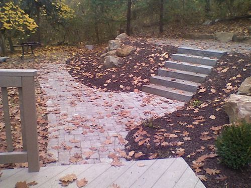 Brick walkway and concrete steps leading up a mulched hill, covered in fallen autumn leaves.
