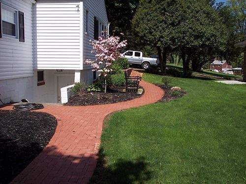 Brick pathway curves around a house with a small tree and green lawn; a truck is parked in the background.