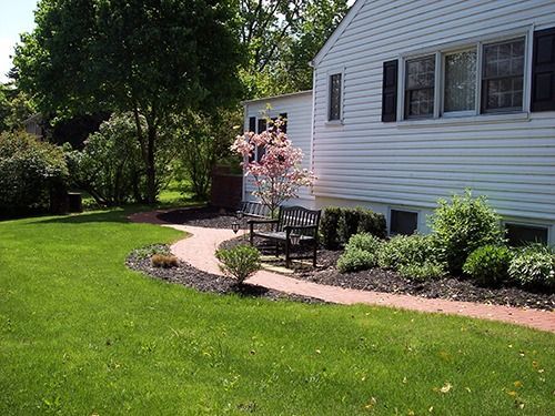 Brick walkway leading to a bench near a white house with black shutters, surrounded by green lawn and shrubs.
