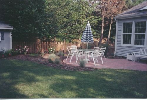 Backyard patio with table, chairs, and umbrella. Surrounded by grass, plants, fence, and house.