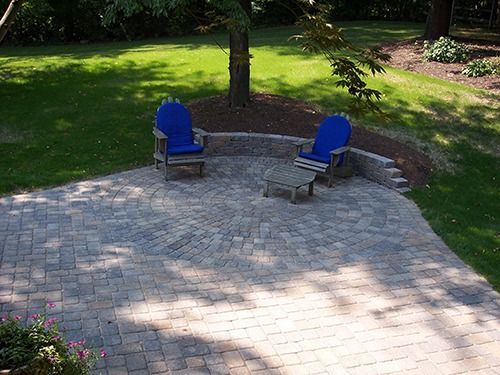 Brick patio with blue Adirondack chairs and a small table under a tree.