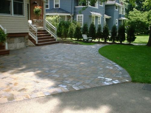 Brick paved driveway, curved edge leading to grass, next to a house with steps.