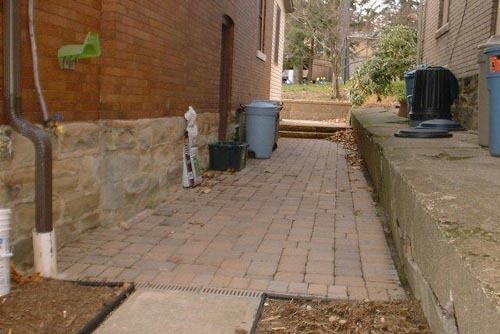 Brick walkway between two buildings; trash cans, a downspout, and a green object on a brick wall.