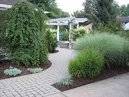 Brick pathway through a landscaped garden with a pergola in the background. Lush greenery and ornamental grasses frame the path.