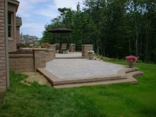 Stone patio with steps and seating area, beside a lawn and trees.