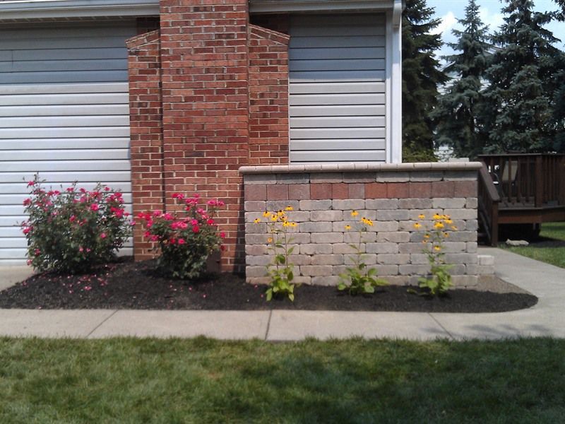 Brick chimney and decorative brick wall with flower beds and bushes along a concrete sidewalk.
