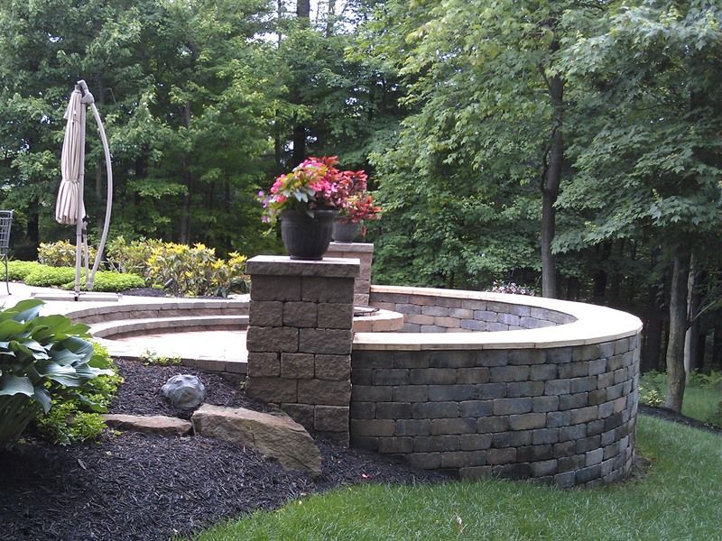 Curved stone retaining wall with potted flowers on top, surrounded by trees and lawn.