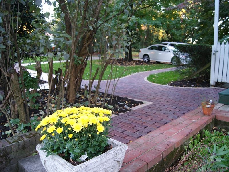 Yellow flowers in a stone planter on a brick path leading to a white car on the lawn.