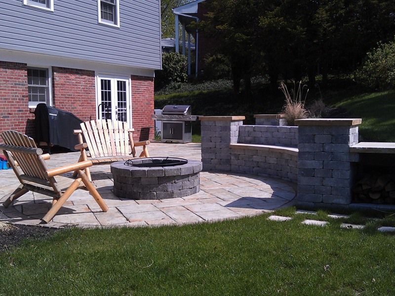 Backyard patio with fire pit, seating, grill, and grass. Brick house in background.