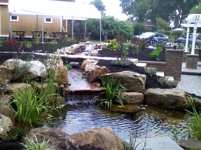 Landscaped water feature with waterfall, pond, and surrounding greenery, near a building and patio.
