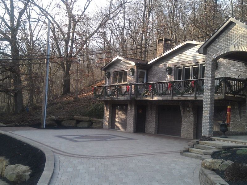 Two-story brick house with garage and balcony, set on a paved driveway in a wooded area.