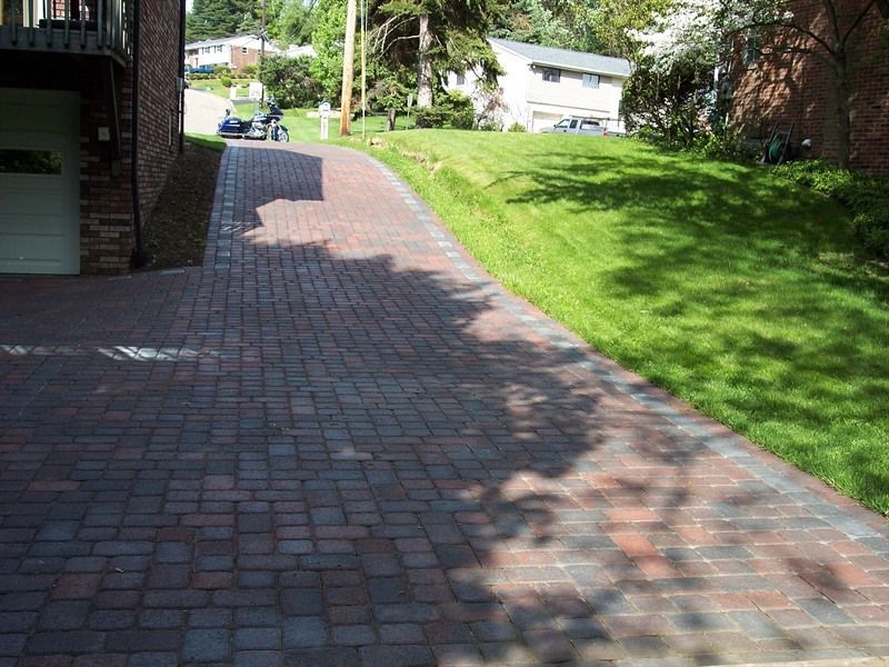 Brick driveway leading uphill, bordered by grass and a building on the left.