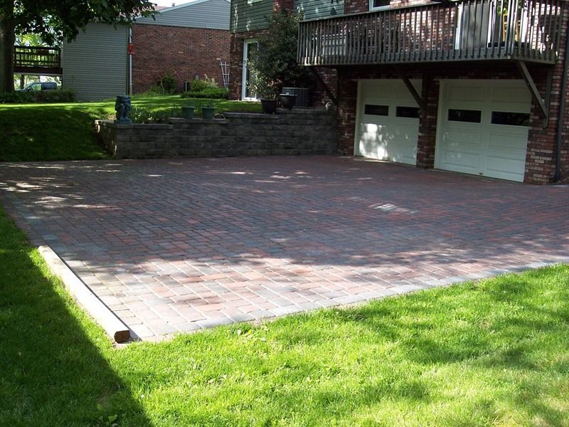 Brick driveway next to a lawn, leading to a two-car garage with a deck above.