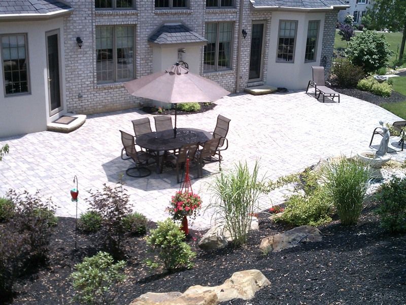 Patio with round table, chairs, and umbrella; surrounded by landscaping and brick house.