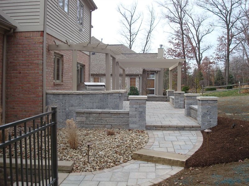 Stone patio with pergola attached to a brick house, pathway leading to it.