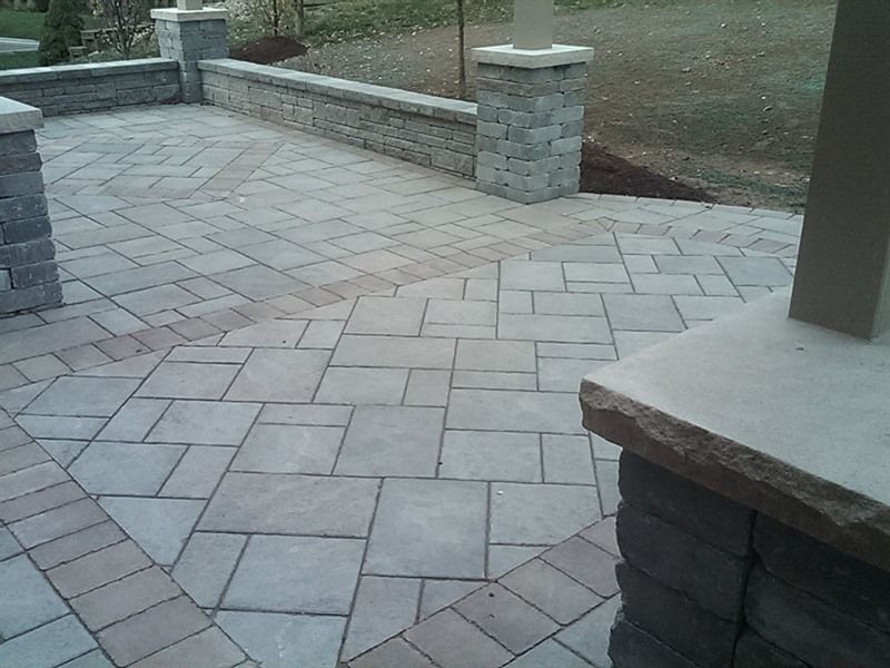 Stone patio with textured pavers, bordered by a low stone wall, stone pillars, and lawn.