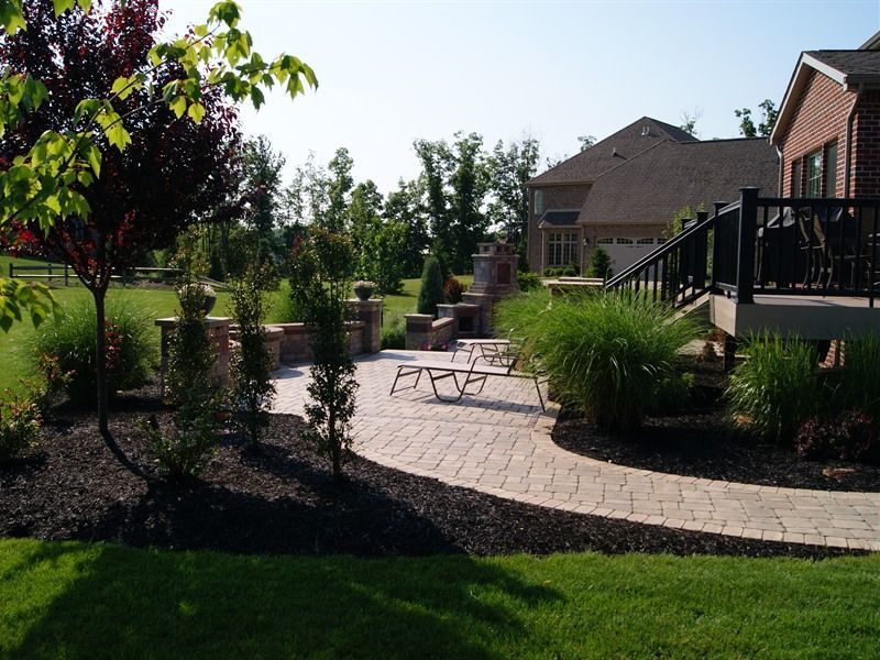 Patio with brick pathway, landscaping, and a house in the background on a sunny day.