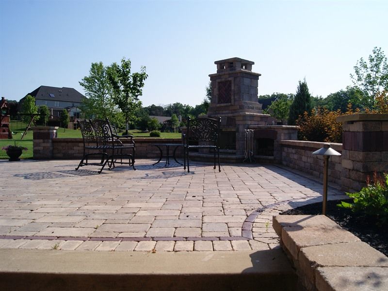 Patio with brick pavers, fireplace, seating, and landscaping under a clear sky.