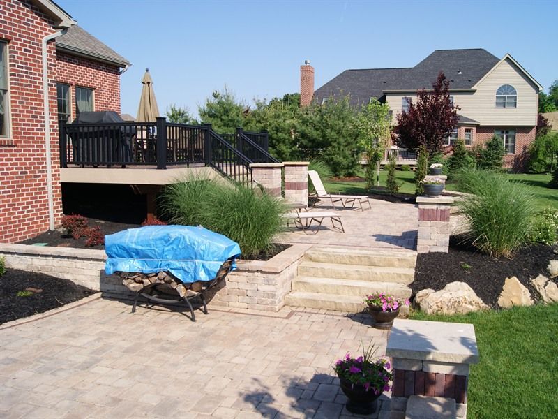 Backyard patio with deck, brick pillars, stairs, landscaping, and a blue-covered object.