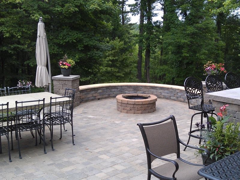 Patio with fire pit, dining table, chairs, and trees in background.