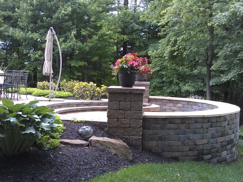 Curved stone retaining wall in a yard, with patio, flowerpot, umbrella, and trees.