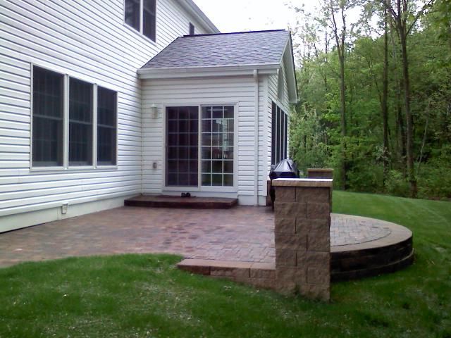 Backyard patio with a stone wall and circular seating area, beside a house with sliding doors and windows.