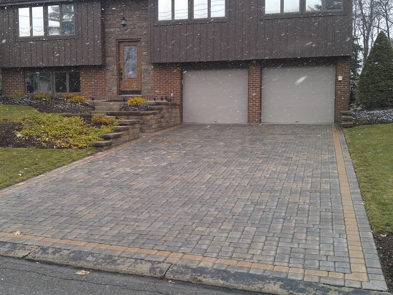 Brick paver driveway leading to a two-car garage under a brick and wood-sided house. Snow is falling.