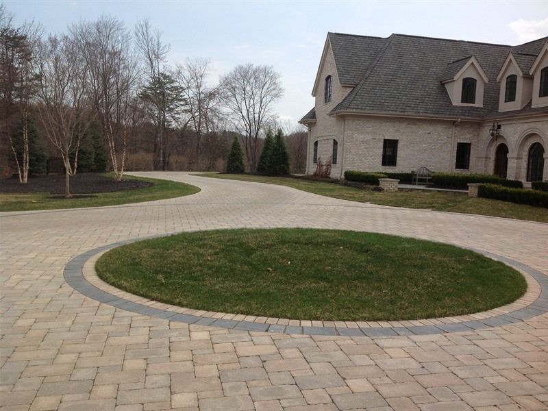 Brick driveway with grass circle in the center, leading to a large house with dormers.