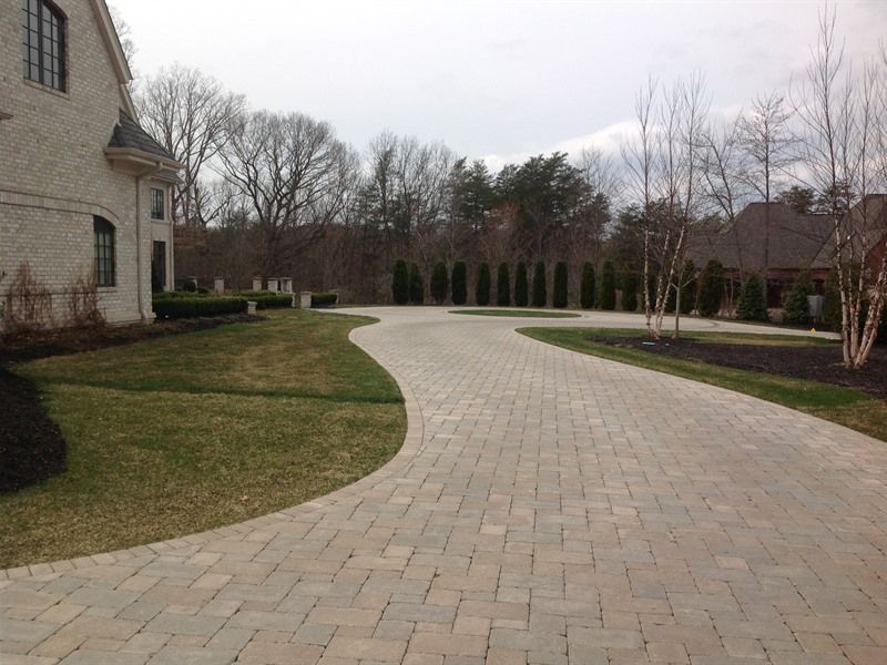 Brick driveway curves through a manicured lawn, leading towards a home and trees in the background.