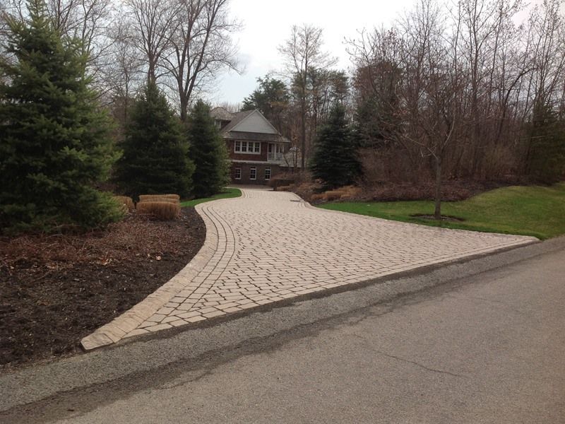 Brick driveway curves towards a large house, surrounded by trees and lawn.