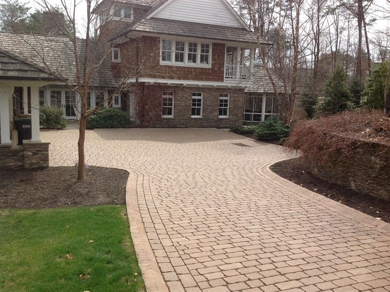 Brick driveway curves toward a multi-story house with wood siding. Green grass and trees frame the scene.