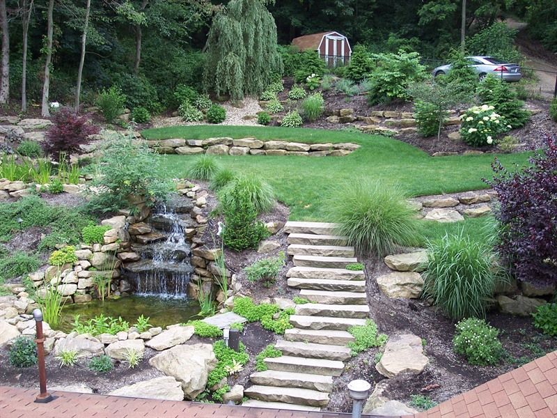 Stone steps lead up a terraced backyard with a waterfall, pond, lawn, and various plants.