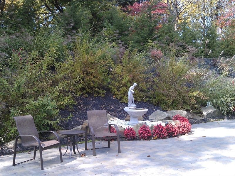 Two chairs face a fountain with a statue. Bushes and colorful flowers surround it on a patio.