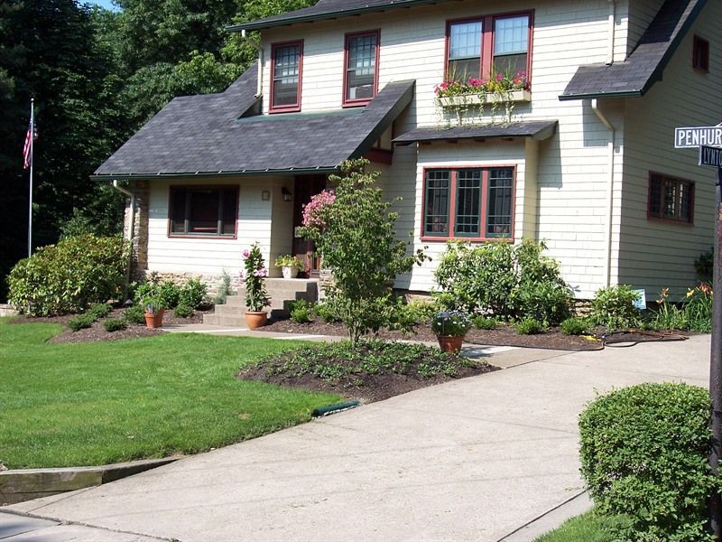Yellow house with brown trim and dark roof, green lawn, trees, and a concrete driveway.