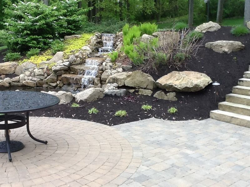 A backyard patio with a waterfall feature, dark mulch, stone steps, and a wrought-iron table.