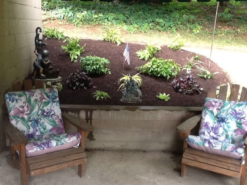 Two wooden chairs with floral cushions on a porch, overlooking a mulch garden and small statues.