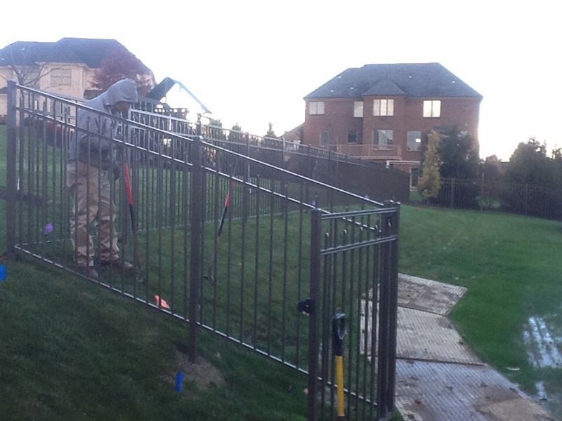 Person installing a black metal fence on a grassy slope, with houses in the background.