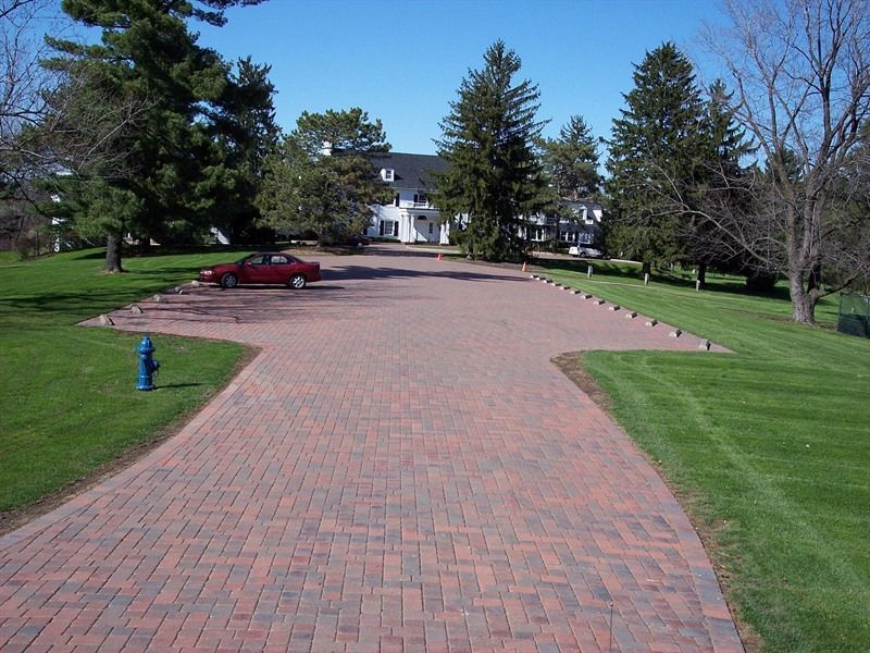Brick driveway leading to a white house, red car parked, blue fire hydrant, green grass, trees, sunny day.