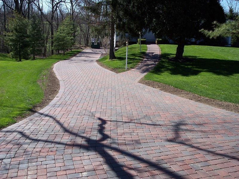 Brick paver driveway splitting into two paths, surrounded by green grass and trees.